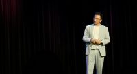 Mann in grauem Anzug spricht gestikulierend auf der Bühne vor Silhouette des Publikums, Quelle: DTF