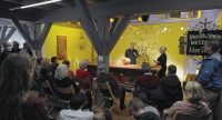 Raum mit Publikum und zwei Personen auf der Bühne, Quelle: YouTube