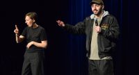 Ein Mann mit Mikrofon spricht und zeigt ins Publikum, neben ihm dolmetscht eine Frau in Gebärdensprache. Dunkler Bühnenhintergrund., Quelle: DTF Stuttgart