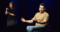 Eine Frau dolmetscht in Gebärdensprache, daneben sitzt ein Mann im Rollstuhl und spricht mit Mikrofon. Beide im Bühnenlicht., Quelle: DTF Stuttgart