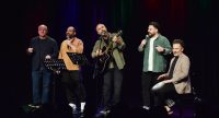 Fünf Männer stehen auf einer Bühne, singen mit Mikrofonen; einer spielt Gitarre, einer sitzt und trommelt. Farbige Publikum im Dunkeln sichtbar., Quelle: DTF Stuttgart