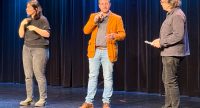 Drei Personen stehen auf einer Bühne: ein Mann spricht ins Mikrofon, links dolmetscht eine Frau in Gebärdensprache, rechts hört ein Mann zu., Quelle: DTF Stuttgart