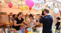 Zwei Frauen stehen hinter einem bunten Marktstand mit Ballons und Geschenken und agieren mit einem Kunden, Quelle: DTF