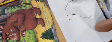 Tisch mit Buntstiften und Grüffelo-Buch. Eine Kinderhand malt ein Bild.