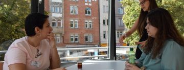 Workshopteilnehmerinnen stehen und sitzen um einen Tisch und sind im Gespräch