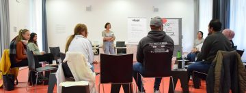 Teilnehmende eines Workshops sitzen im Halbkreis um eine stehende Moderatorin