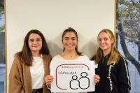 Drei junge Frauen stehen nebeneinander und lächeln, eine hält ein Schild, Quelle: DTF