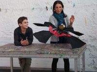 Junge lehnt sich lächelnd an einem Holztisch, junge Frau rechts neben ihm hält einen Vampir aus einem Regenschirm gebastelt, Quelle: DTF