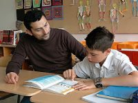 junger Mann und Junge schauen gemeinsam in ein Arbeitsheft, sie sitzen in einem Schulzimmer, Quelle: DTF