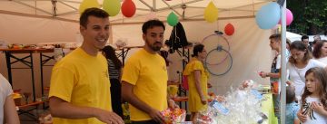 Zwei junge Männer in gelben Shirts stehen hinter einem Marktstand mit Geschenken, Quelle: DTF